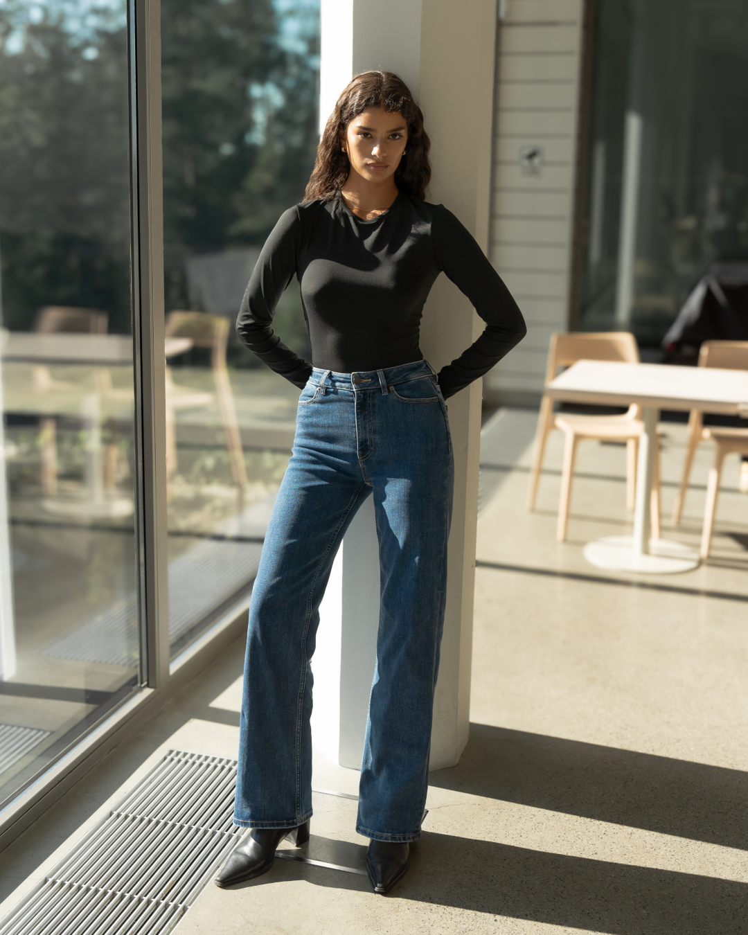 Woman standing by window wearing mid blue Ninepine Relaxed ComfortDenim jeans with a black Soft Long Sleeve T-Shirt and black boots.