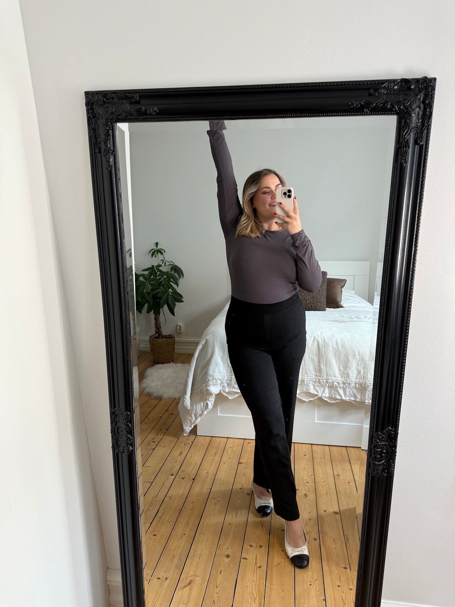Woman taking a mirror selfie wearing black tailored straight trousers with a fitted long-sleeve soft top, styled with two-tone flats for a clean, minimal everyday outfit