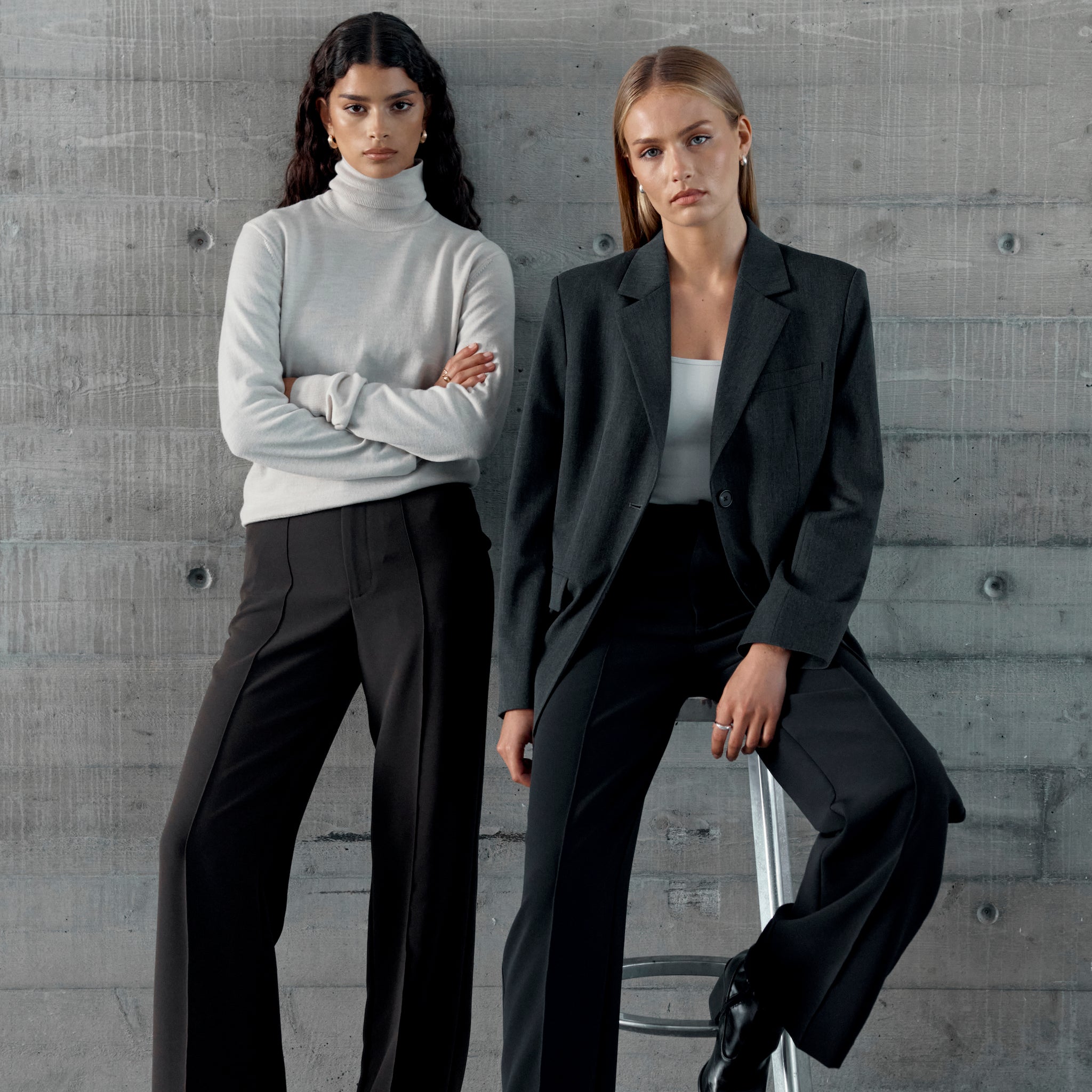Two women wearing Ninepine office trousers styled for work—one in a light turtleneck with dark trousers, the other in a blazer with black tailored trousers—posed against a concrete wall.